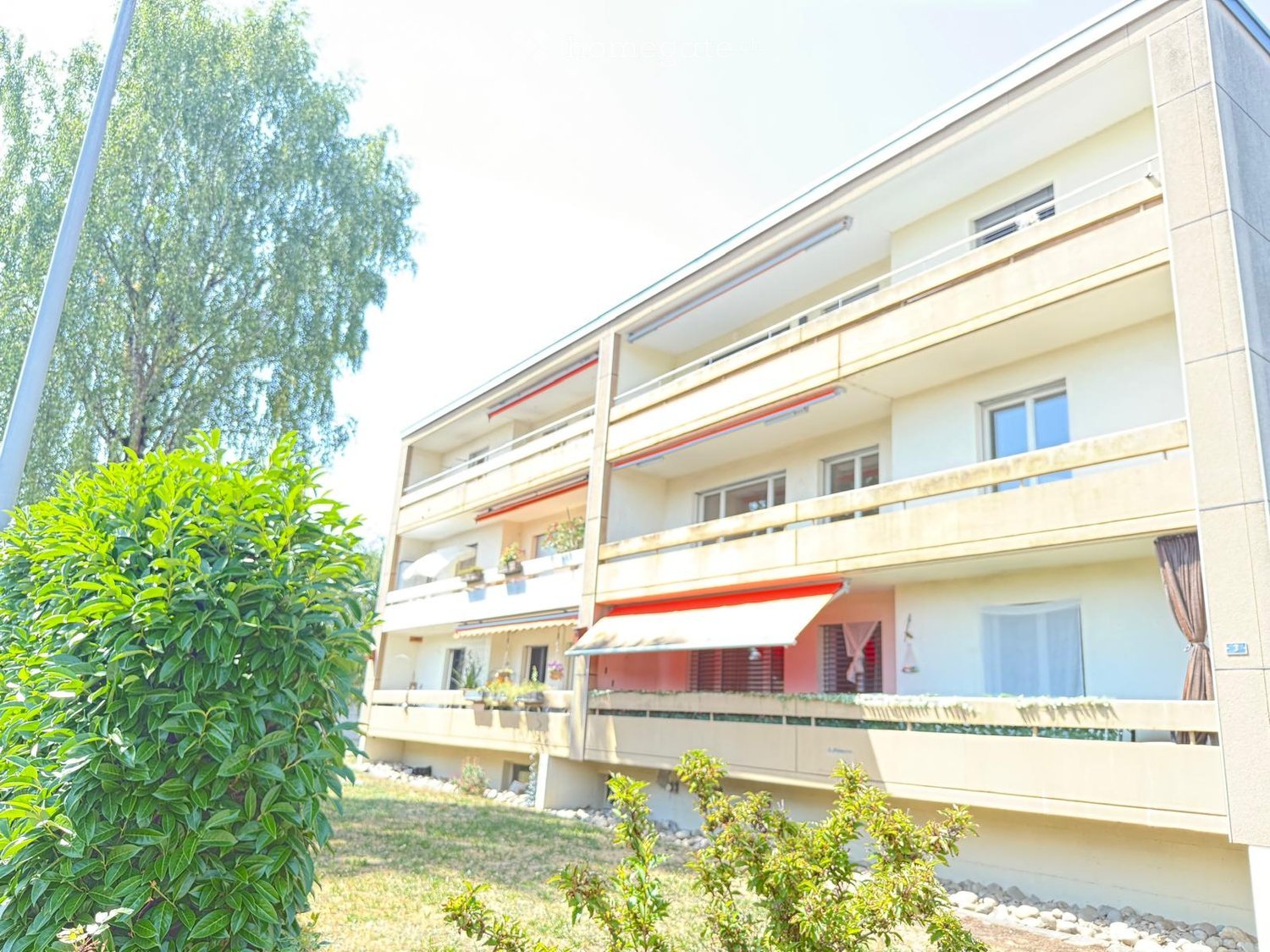 A multi-story residential building with balconies, large windows, and a modern facade. The building is surrounded by greenery and has a well-maintained lawn.