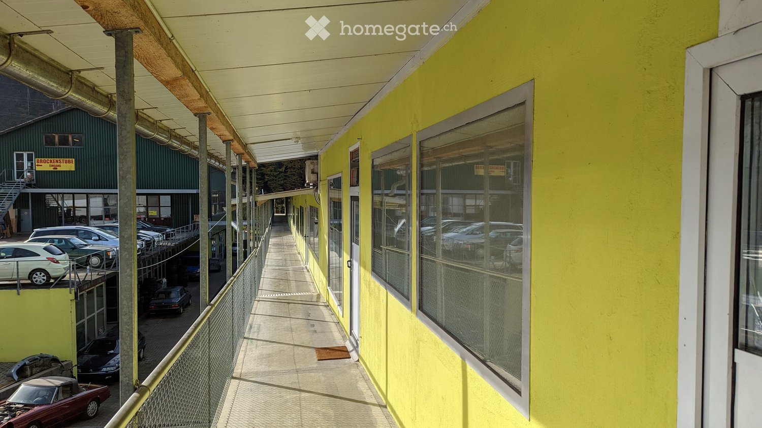 A covered walkway with yellow walls, metal railings, and large windows, leading to a parking area with several cars.