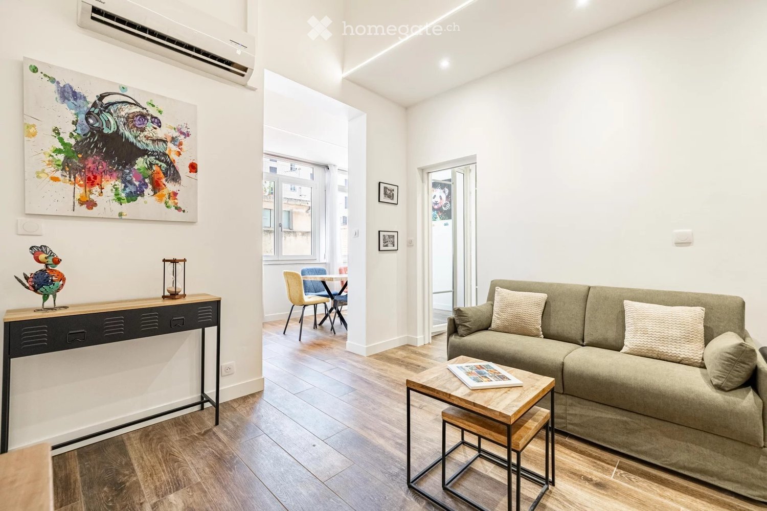 Modern living room with light-colored sofa, coffee table, small wooden stool, dining area with table and chairs, artwork on wall, and decorative sculpture.