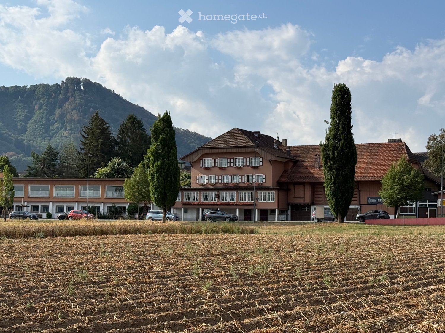 Two-story building with red-tiled roof and multiple windows, surrounded by trees and parked cars, with a grassy field in the foreground and mountains in the background.