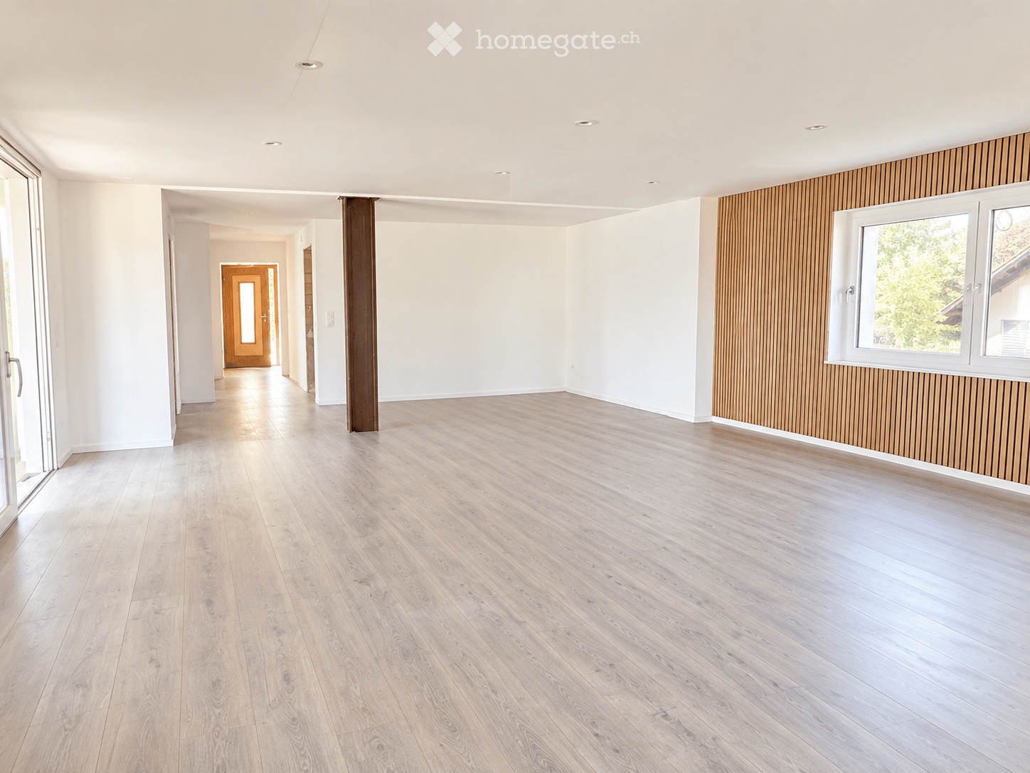 Empty room with light wooden flooring, white walls, and a large window with white frames. The room features a modern design with recessed ceiling lights and a wooden accent wall.