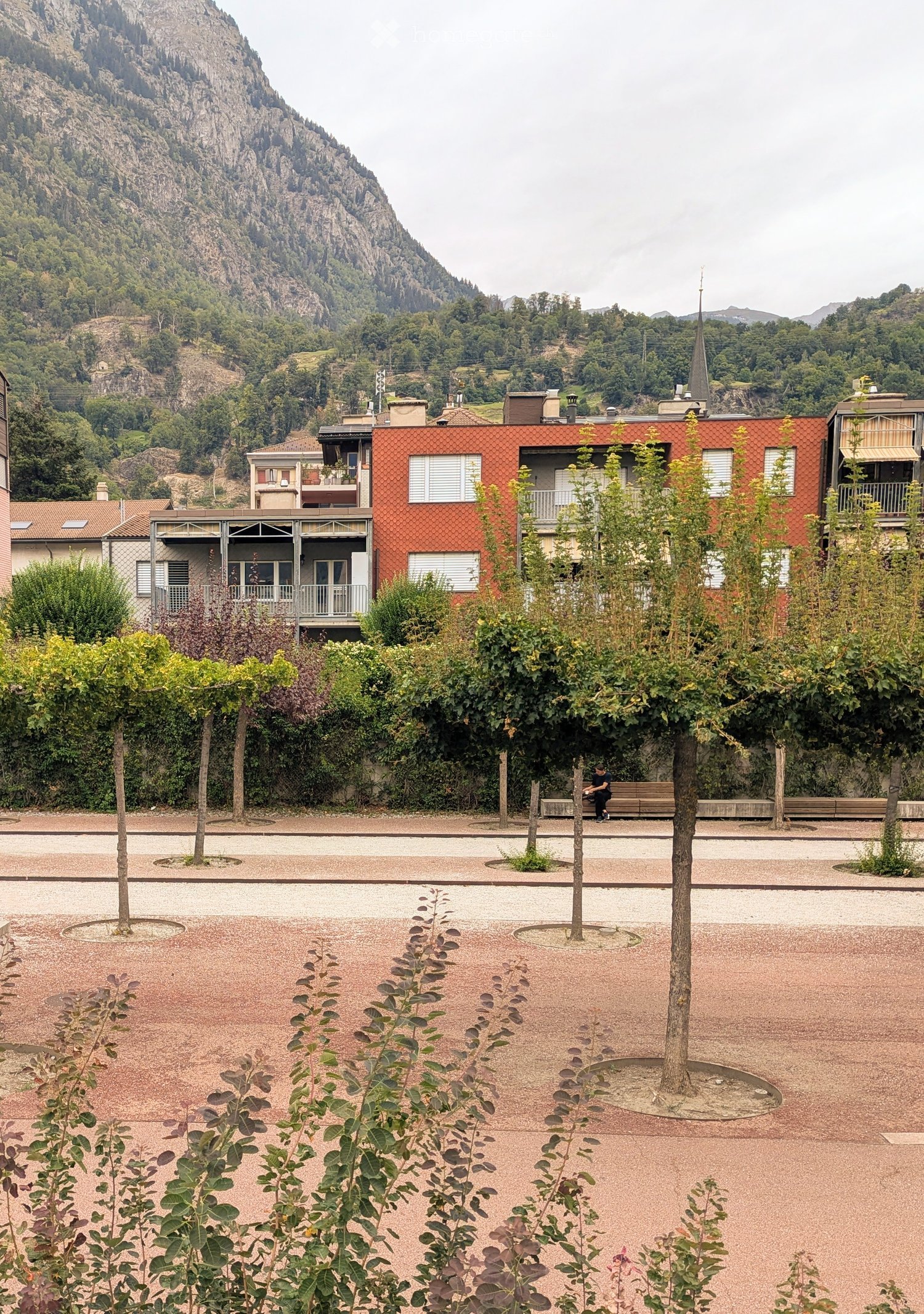 A landscaped outdoor area with trees, benches, and modern buildings in the background, set against a mountainous backdrop.