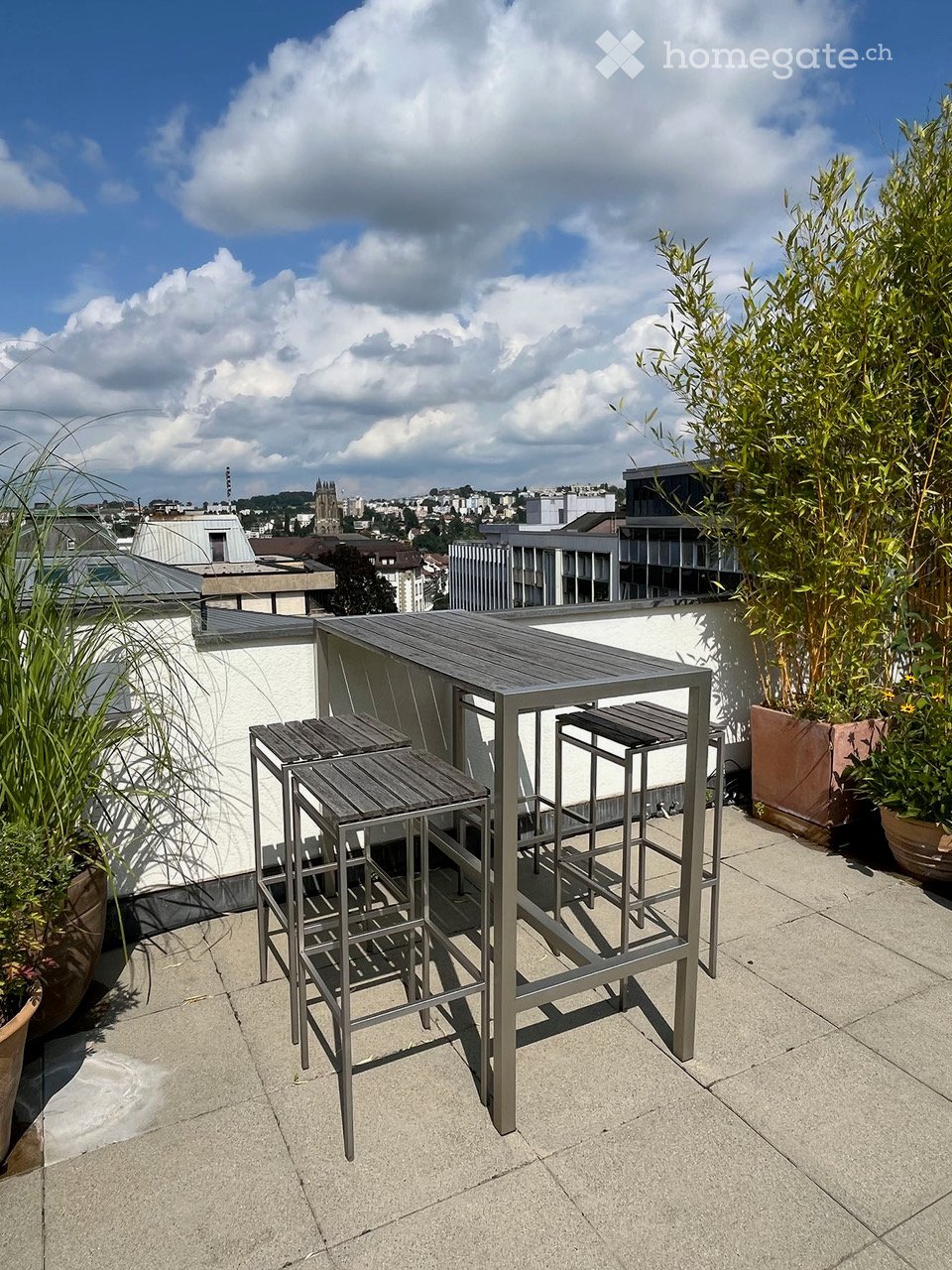 Rooftop terrace with a modern metal table and stools, surrounded by potted plants, offering a cityscape view.