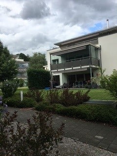 Modern two-story house with white exterior, large windows, balcony, and well-maintained garden.