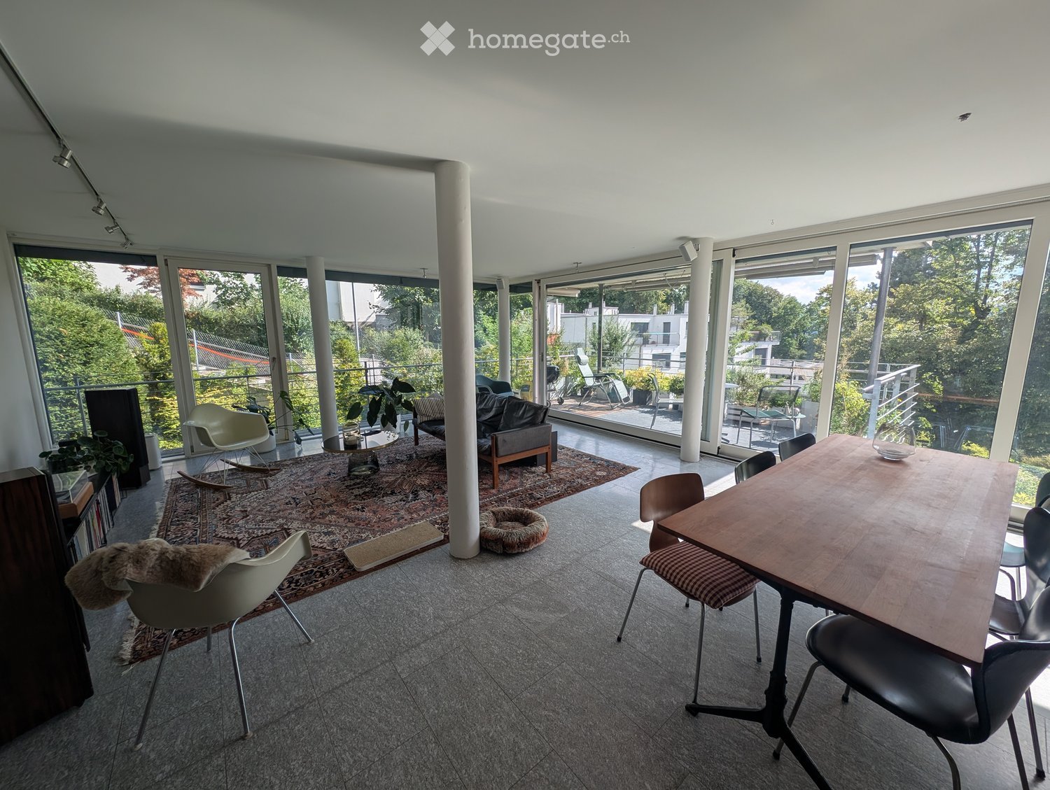Bright living and dining area with large windows and a balcony, featuring a sofa, dining table, chairs, and plants.