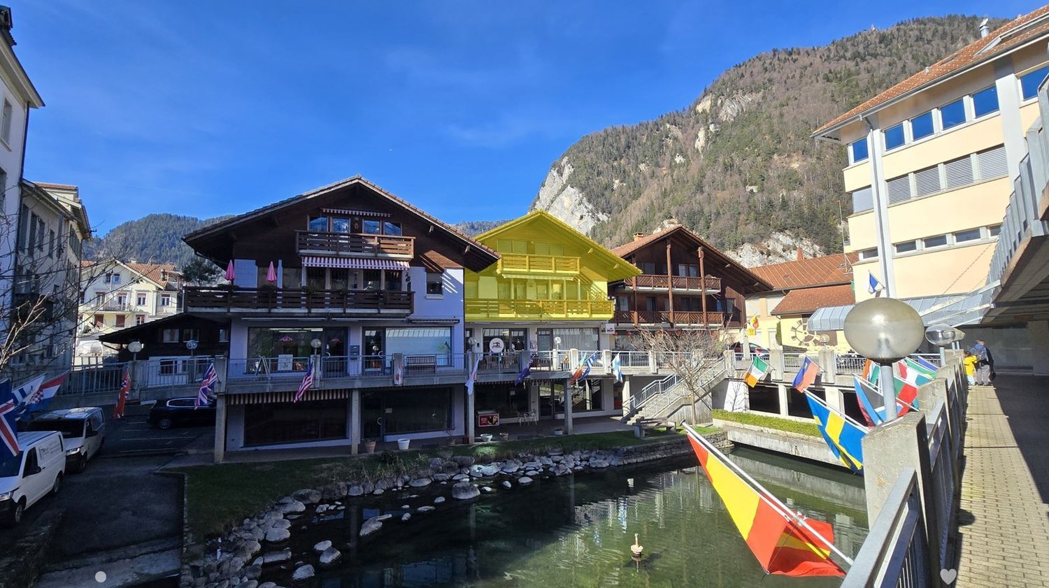 A picturesque town square with colorful buildings, a small canal, and a backdrop of mountains.
