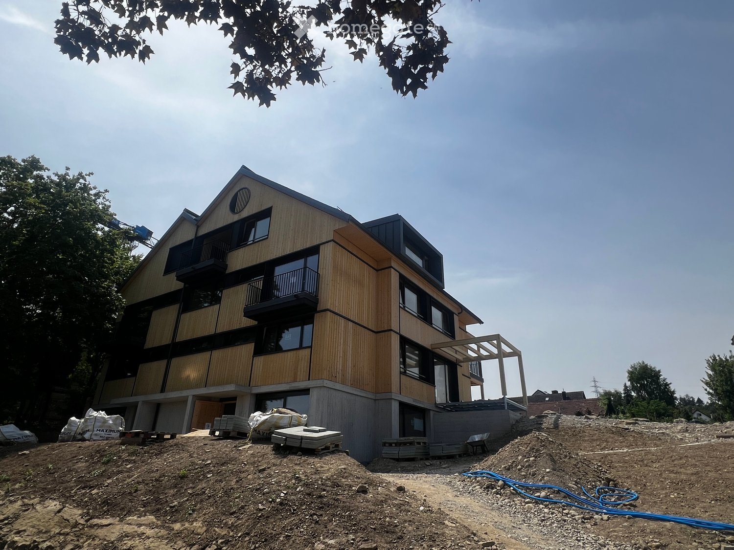 Modern two-story house with wooden siding, large windows, and balconies, surrounded by construction materials and trees.