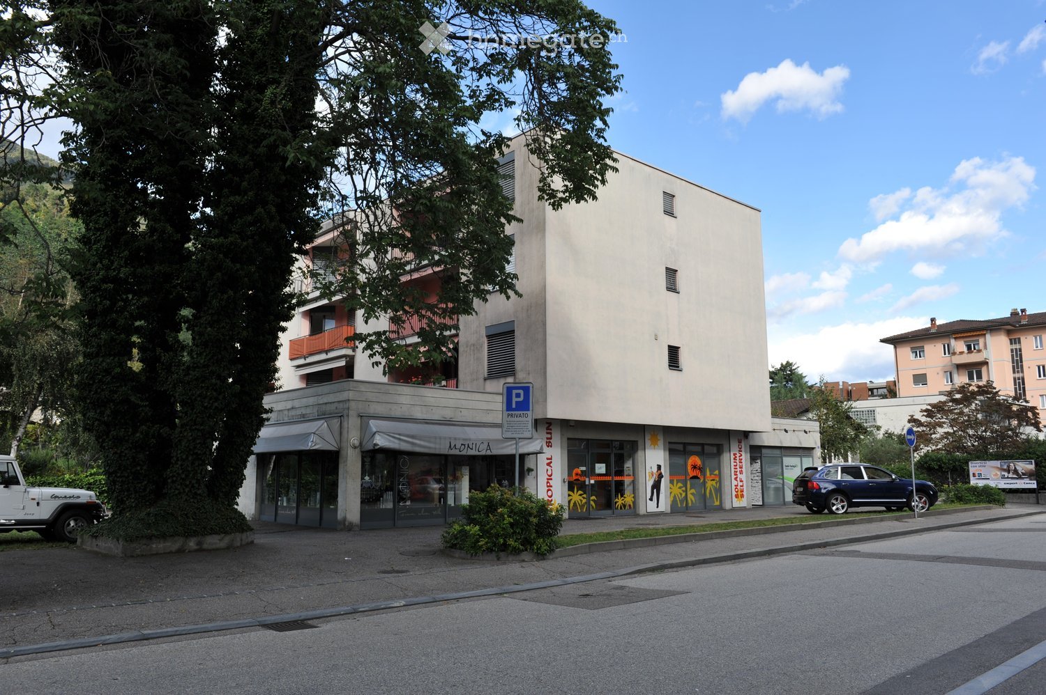 Modern multi-story building with a storefront labeled 'MONICA' and 'OPTICAL SUN' on the ground floor, surrounded by trees and parked cars.