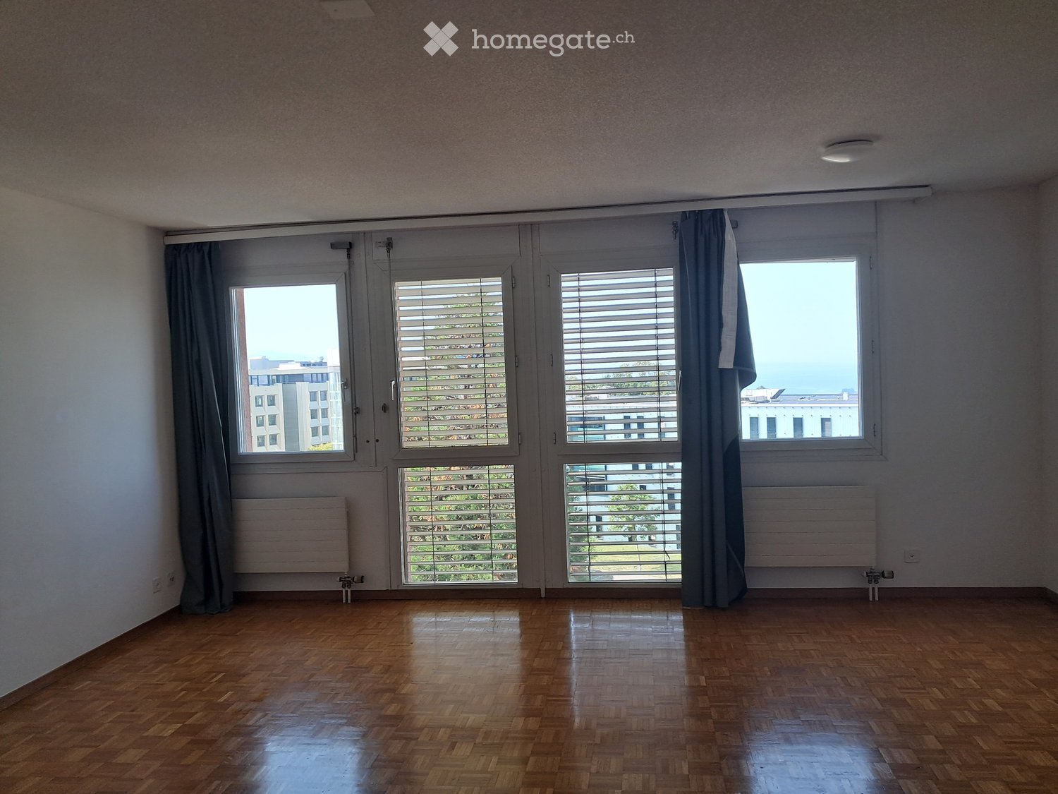 Empty room with wooden flooring, large windows with blinds, and radiators. No furniture or personal items present.
