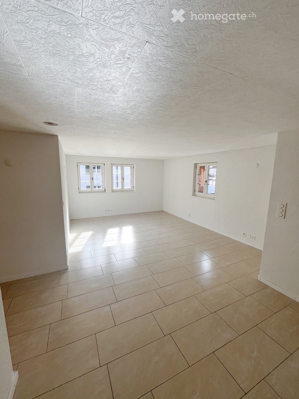 Empty room with light-colored tiled flooring and white walls, featuring three windows and a textured ceiling.