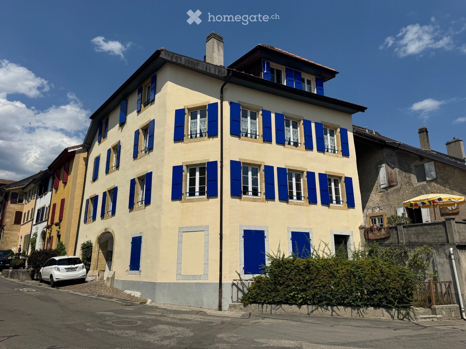 A three-story residential building with light yellow walls, blue shutters, and multiple windows. The building features a chimney and is situated on a street with a parked car and neighboring houses.