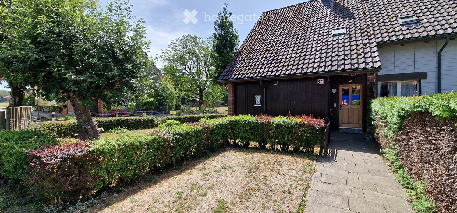Single-story house with a dark wooden facade, tiled roof, and a small garden area with trimmed hedges and a tree.