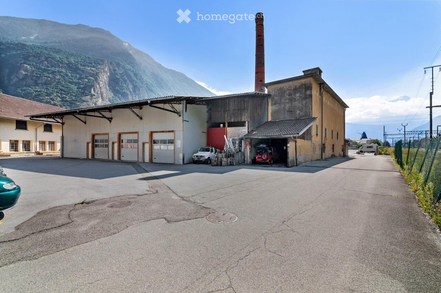 Industrial building with large garage doors, a tall chimney, and a mountainous backdrop. Vehicles are parked in front, and the area includes a paved driveway.