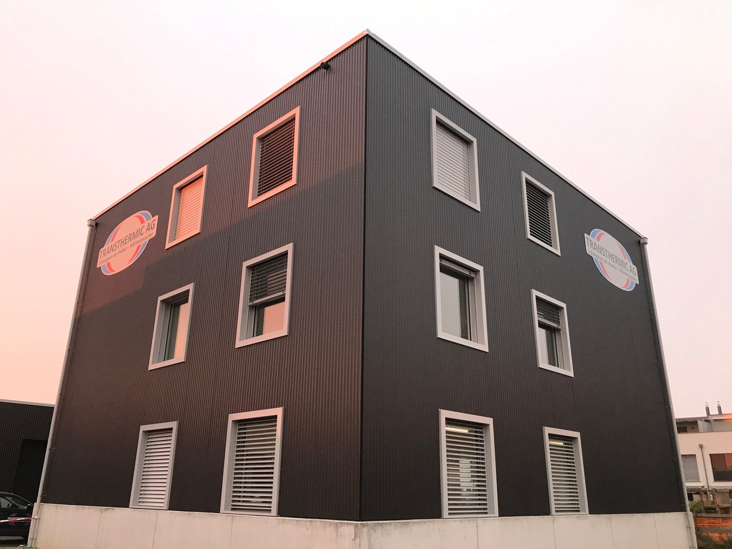Modern two-story building with dark brown metal siding, multiple windows with white frames, and company logos on the facade.