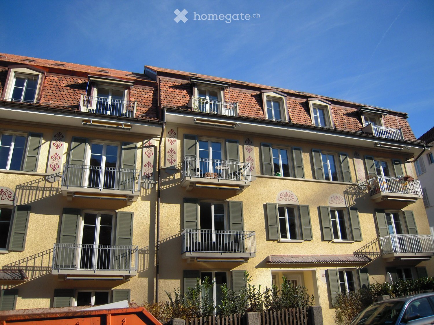Multi-story residential building with yellow facade, green shutters, multiple balconies, and a red-tiled roof.