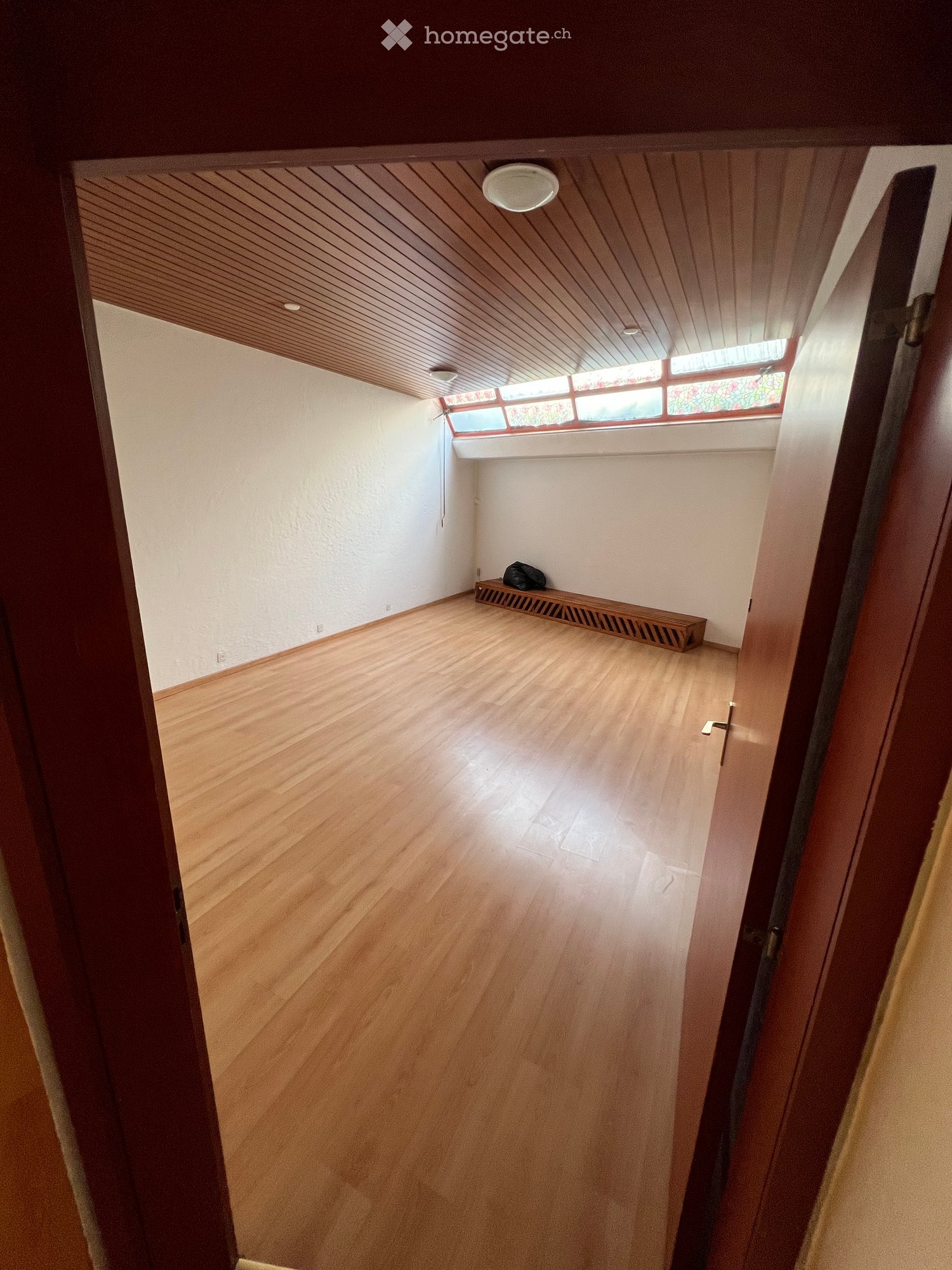 Empty hallway with wooden flooring, white walls, and a slanted wooden ceiling. A small bench and a bag are visible against one wall.