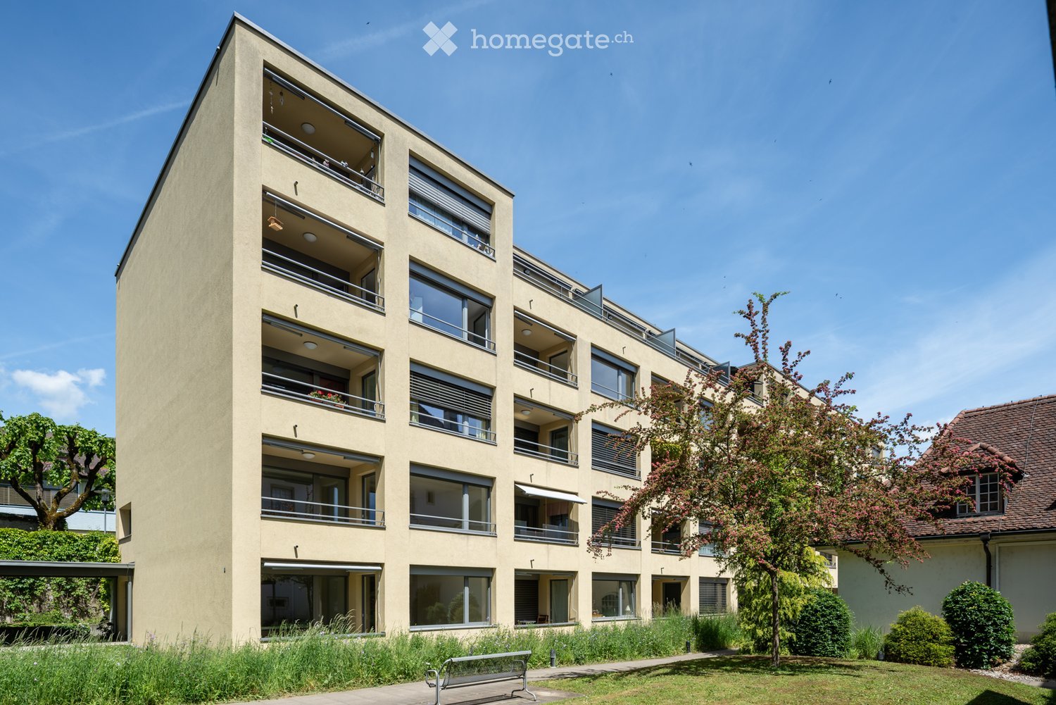 Modern multi-story residential building with balconies, large windows, and a landscaped garden area in front.