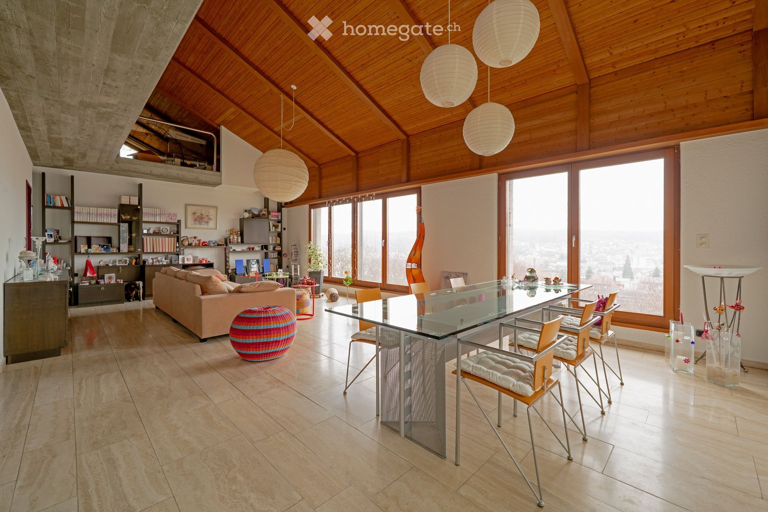 A spacious living room with a high, wooden-beamed ceiling, large windows offering a view, a beige sofa, a colorful ball, a glass dining table with chairs, shelves with books and decorative items, and pendant lamps.