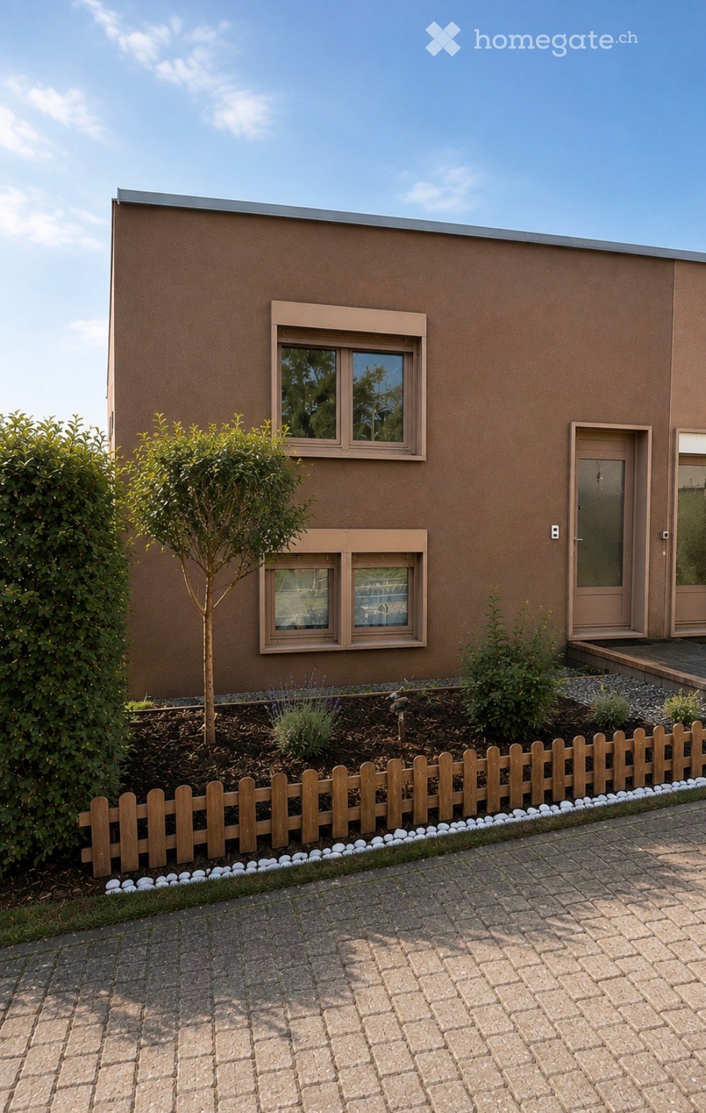 Single-story house with brown facade, white-framed windows, and a small garden area with a wooden fence and decorative white pebbles.