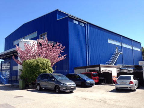 A large industrial-style building with a blue exterior and white trim, featuring a garage and parking area with several cars.