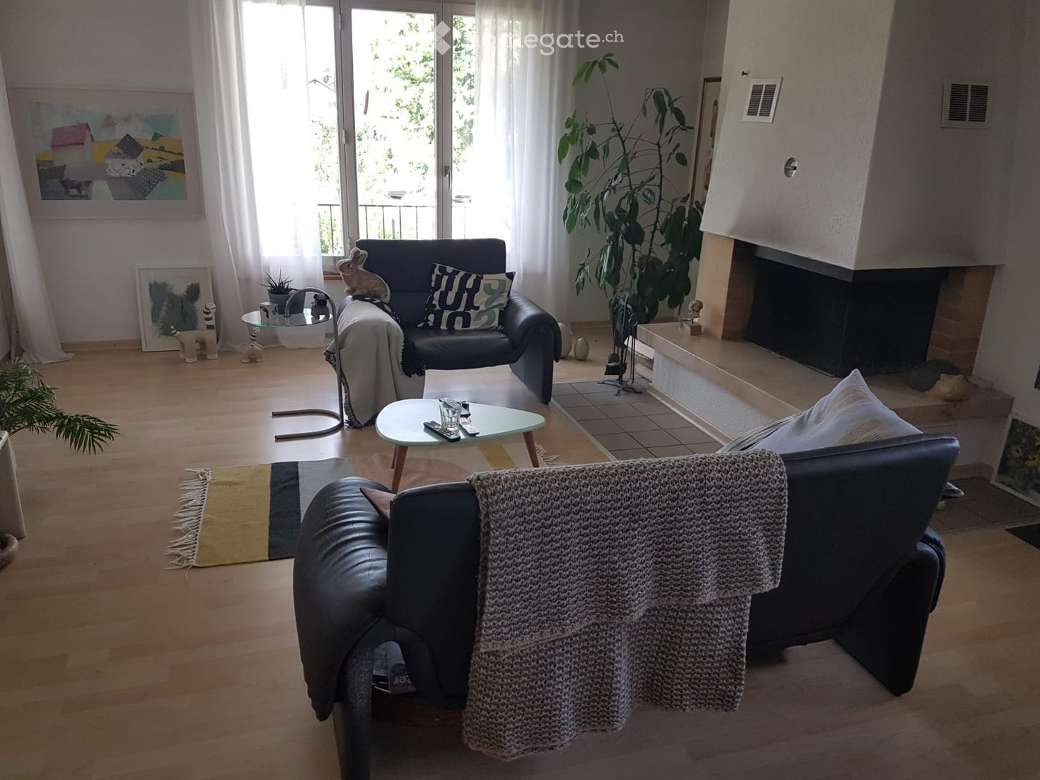 Modern living room with hardwood floors, fireplace, large windows, balcony access, sofa, coffee table, armchairs, ottoman, floor lamp, and decorative items.