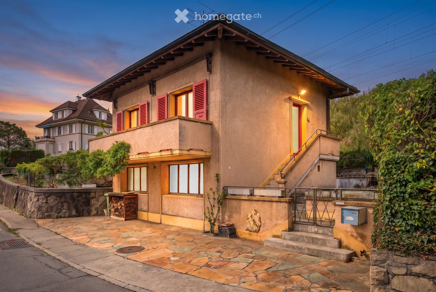 Two-story house with beige facade, red shutters, and a raised ground floor accessed by stairs. Features a stone-paved entrance area, small garden, and a balcony on the upper level.