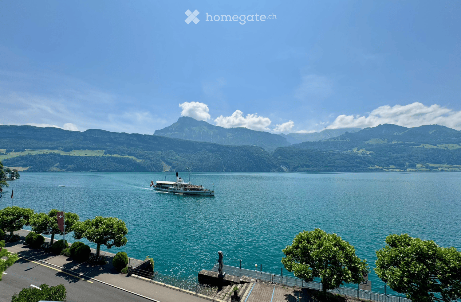 A scenic view of a lake with a boat sailing on it, surrounded by lush green trees and distant mountains under a clear blue sky.