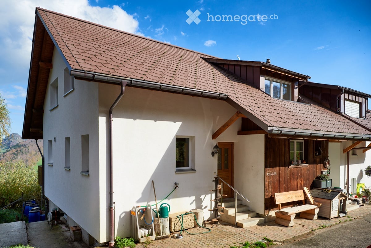Two-story house with white walls, brown roof, wooden door, and small porch area with bench and tools.