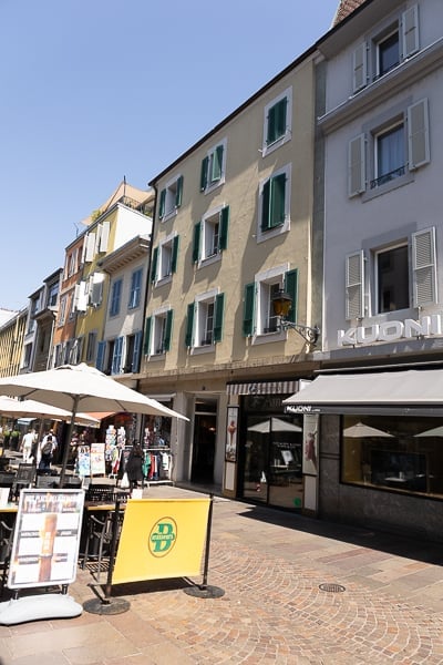 A charming outdoor area with a cobblestone pavement, colorful buildings with shutters, and outdoor seating under umbrellas.