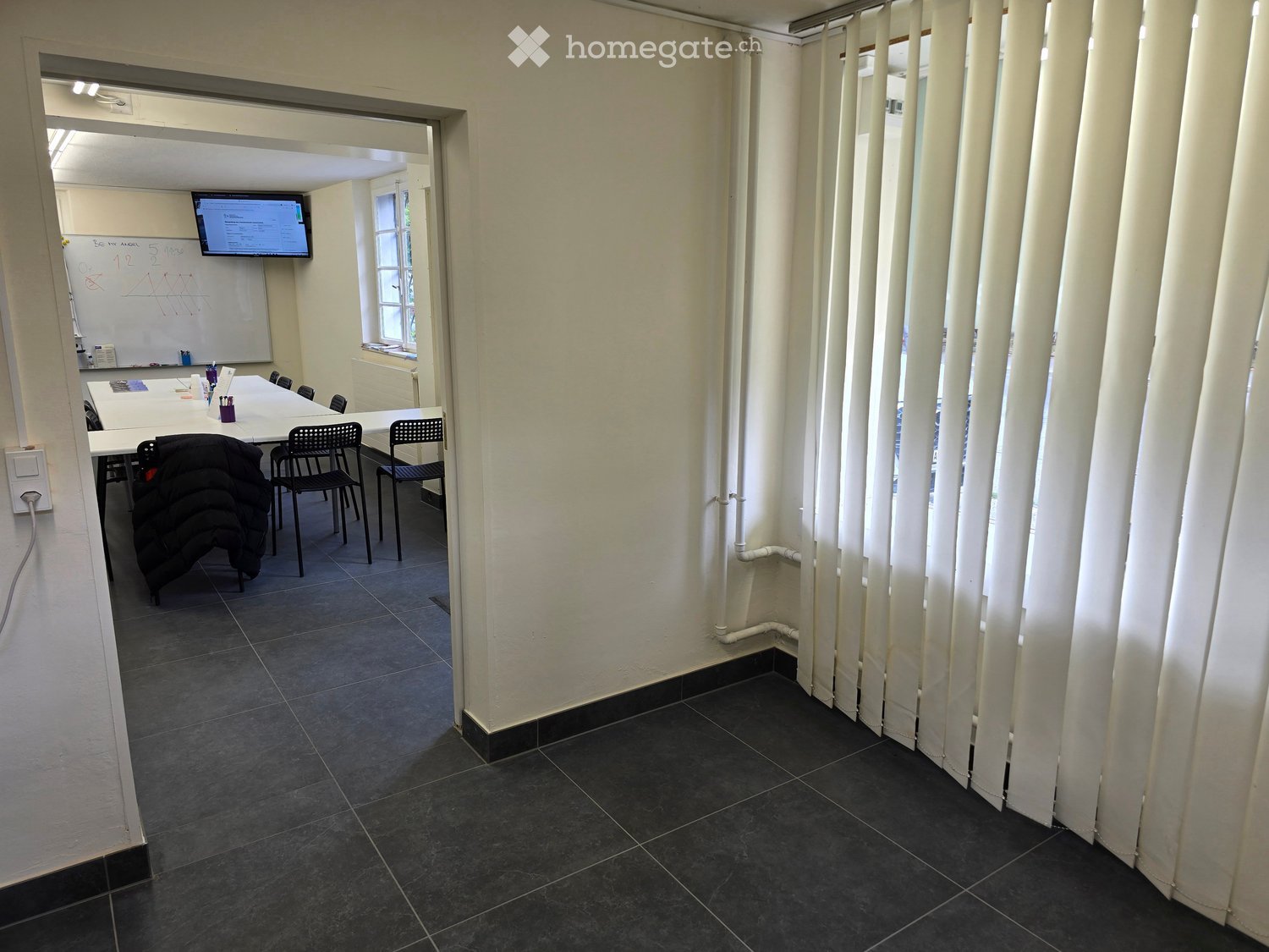A furnished room with a table, chairs, a screen, and a whiteboard. The room has white walls, dark tiled flooring, and vertical blinds on one side.