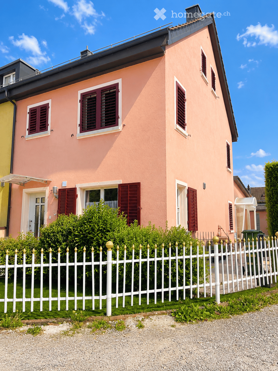 Pink two-story house with white-framed windows and red shutters, surrounded by a white picket fence and greenery.