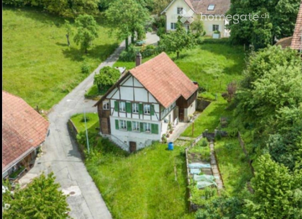 Aerial view of a traditional two-story house with a red-tiled roof and green shutters, surrounded by a well-maintained garden and trees.