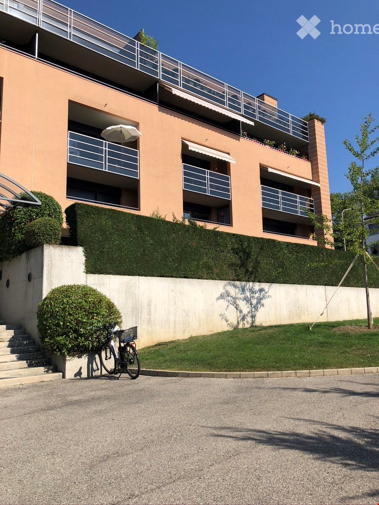 Modern multi-story building with balconies, light pink facade, and neatly trimmed hedges. A bicycle is parked near the entrance.