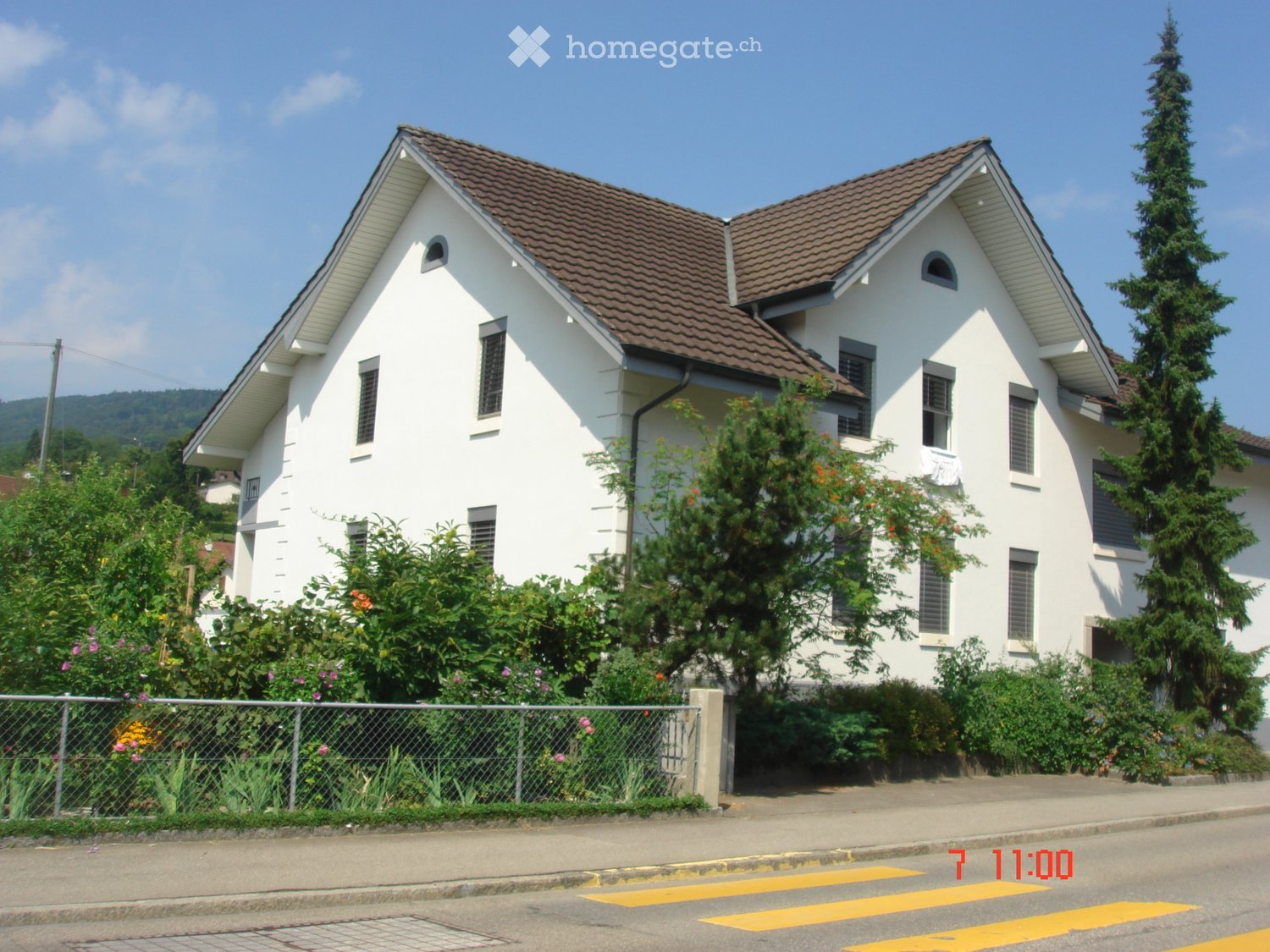 Two-story white house with brown tiled roof, multiple windows, and surrounding greenery.