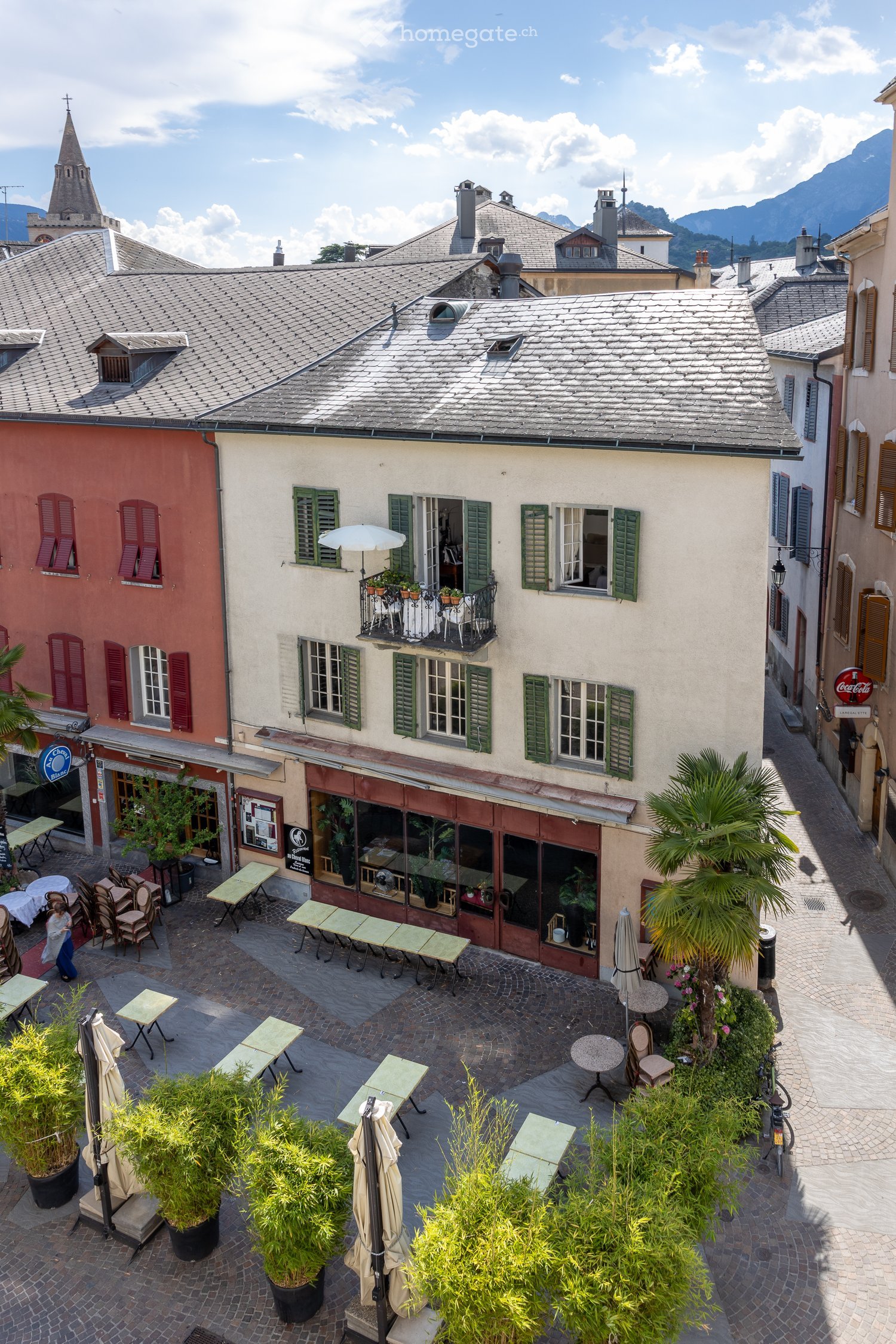 Courtyard with outdoor seating, tables, chairs, potted plants, and a small balcony with greenery and an umbrella.