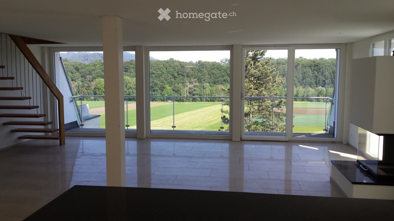 Bright, open living space with large glass doors leading to a balcony, wooden staircase, and tiled floor.