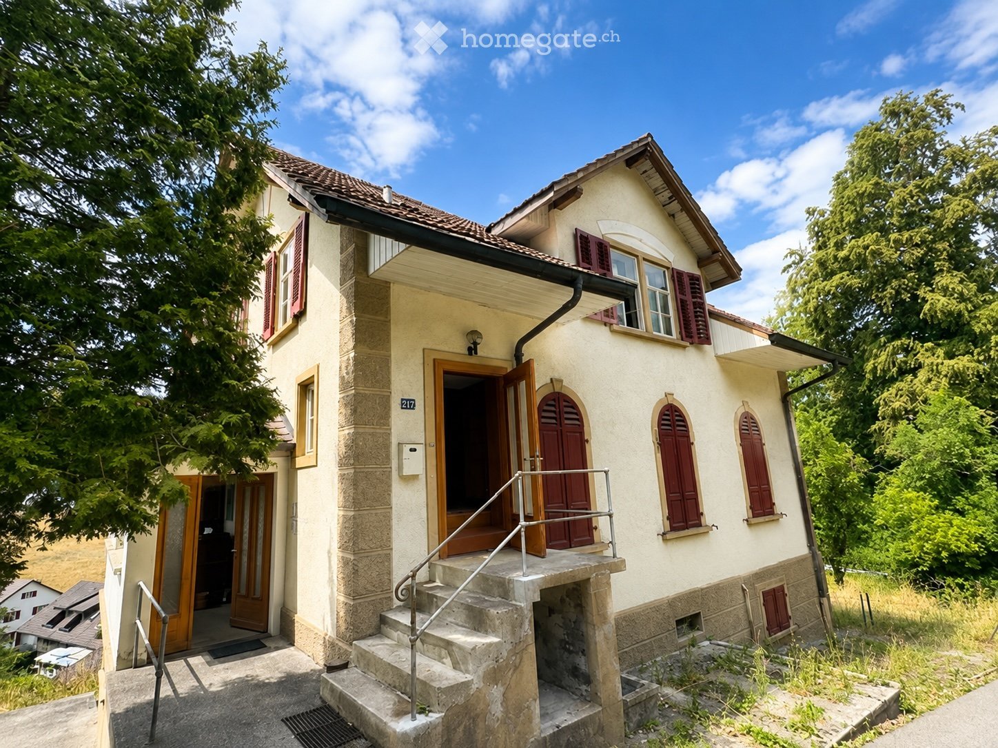 Two-story house with cream-colored walls, red shutters, and a tiled roof. Features a stone staircase leading to the entrance, with a small porch and a raised ground floor.