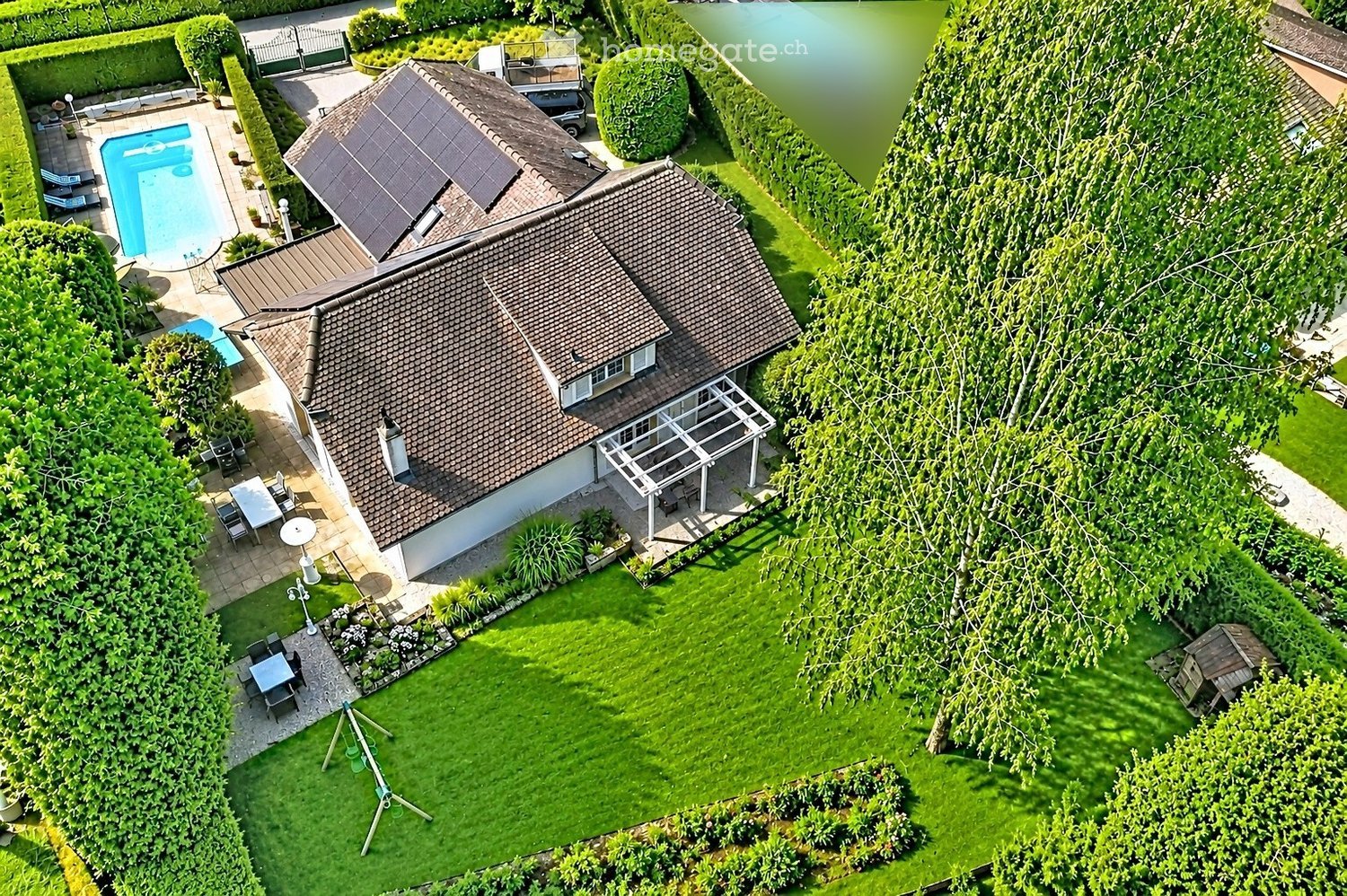 Aerial view of a house with a well-maintained garden, swimming pool, patio furniture, and a covered outdoor seating area.
