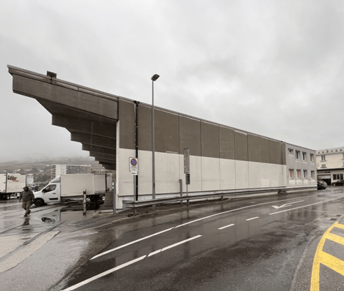 Modern industrial-style building with a large, angular concrete facade and a prominent overhanging roof. The building is situated near a road with visible traffic signs and a few parked vehicles, including a white van. The scene appears to be on a rainy d