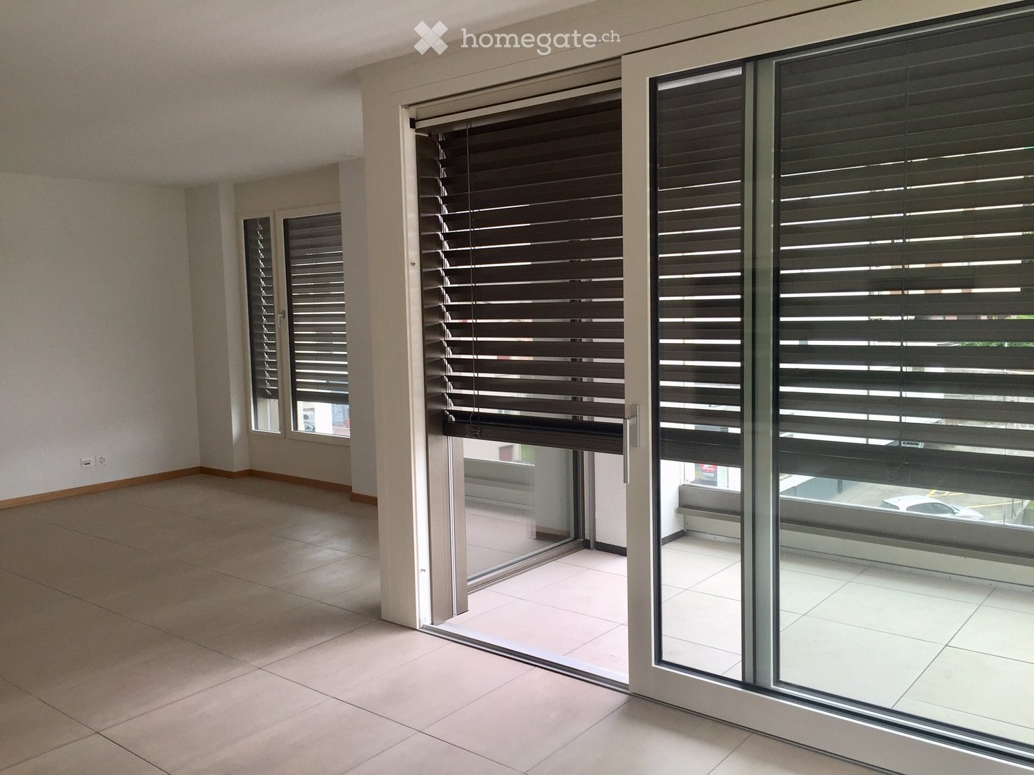 Empty room with tiled floor, large windows with blinds, and a sliding glass door leading to a balcony area.