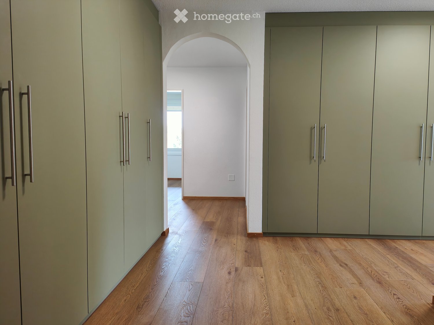 A modern hallway with light green cabinetry, wooden flooring, and an arched opening leading to another room.