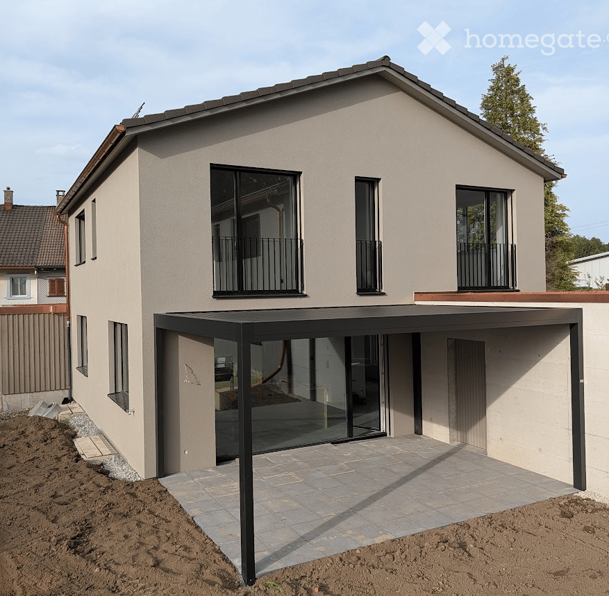 Modern two-story house with beige facade, large windows, and balconies. Features a covered patio area with glass walls and a tiled floor.