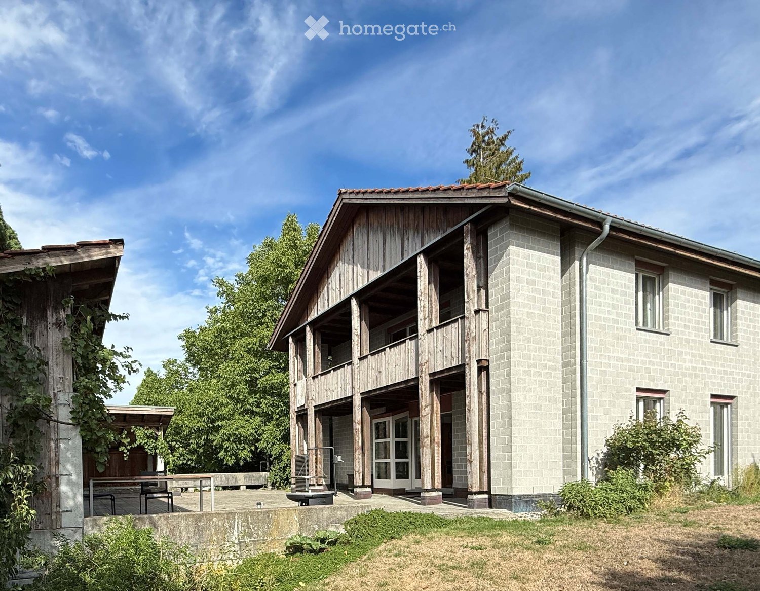 Two-story house with wooden balcony and stone facade, surrounded by greenery and a raised ground floor.