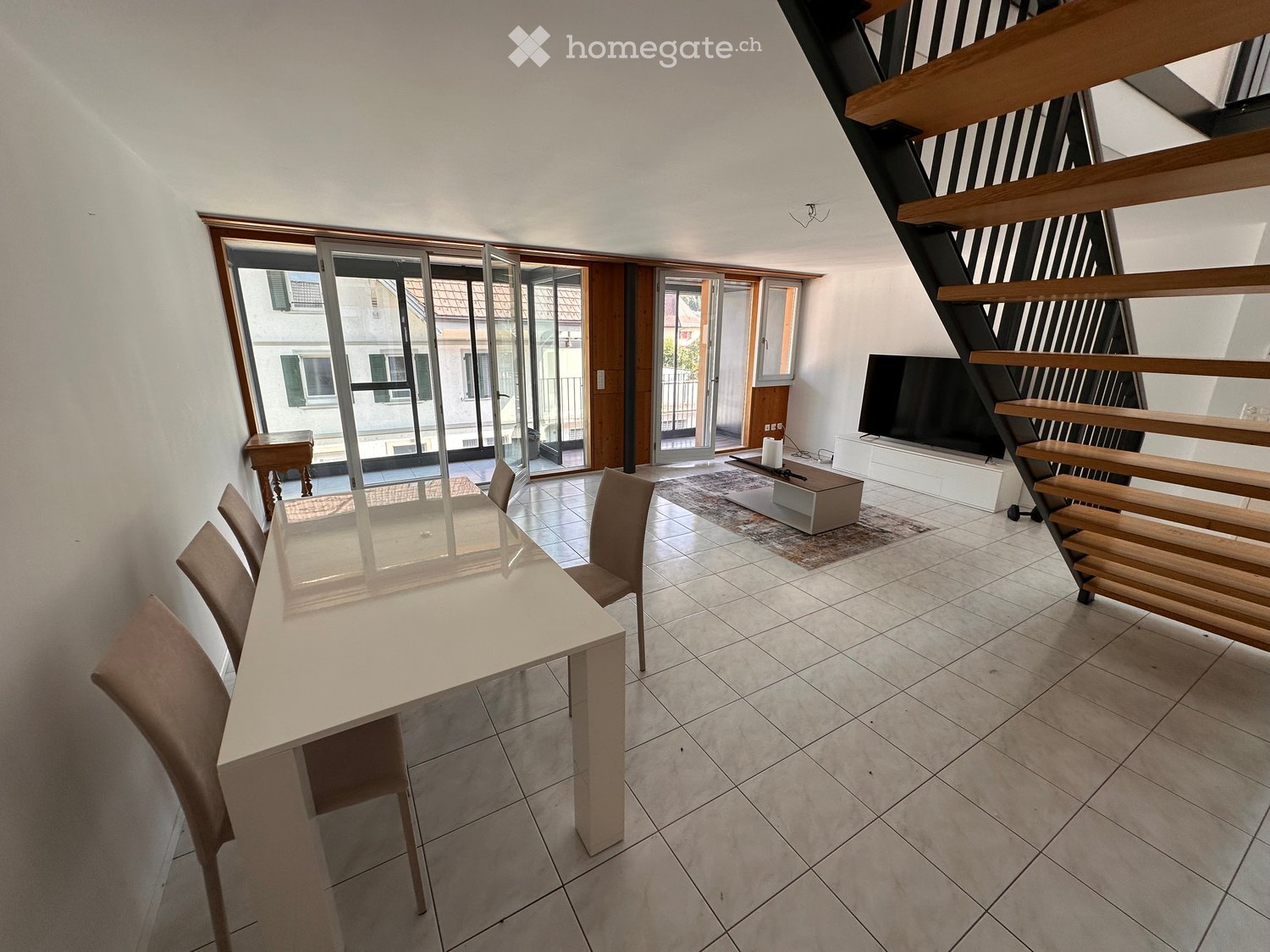 Bright, modern dining area with a white dining table, four chairs, a television on a stand, and a small wooden table near the sliding glass doors leading to a balcony.