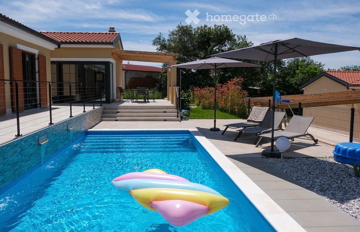 Outdoor area with a swimming pool, lounge chairs, umbrella, and a modern house in the background.