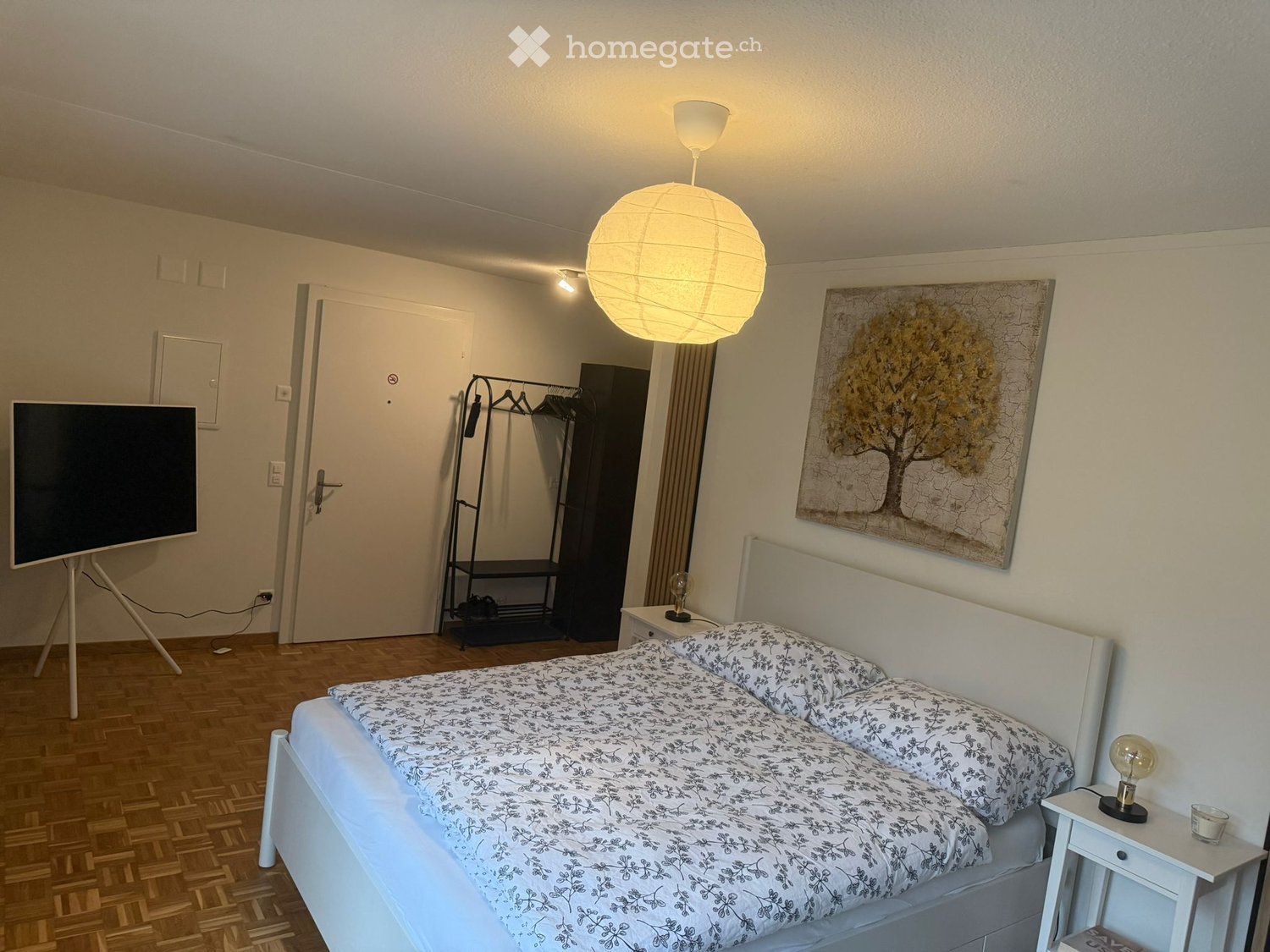 A modern bedroom with a double bed, nightstand, lamp, painting, clothing rack, and a large screen. The room features wooden flooring and white walls.