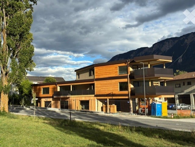 Modern multi-story building with wooden facade, balconies, and large windows, situated in a scenic area with mountains in the background.