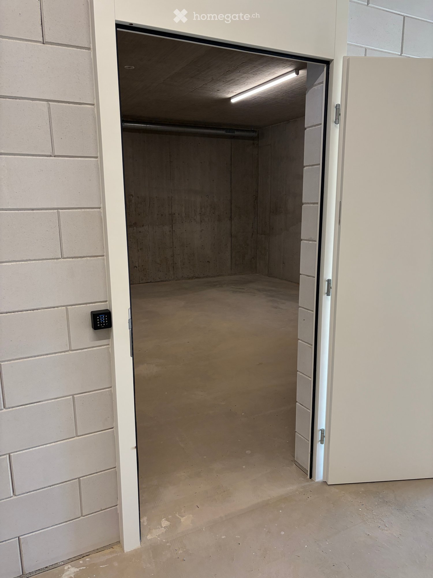 Empty garage with concrete floor, white brick walls, and an open door leading to a concrete-floored space.