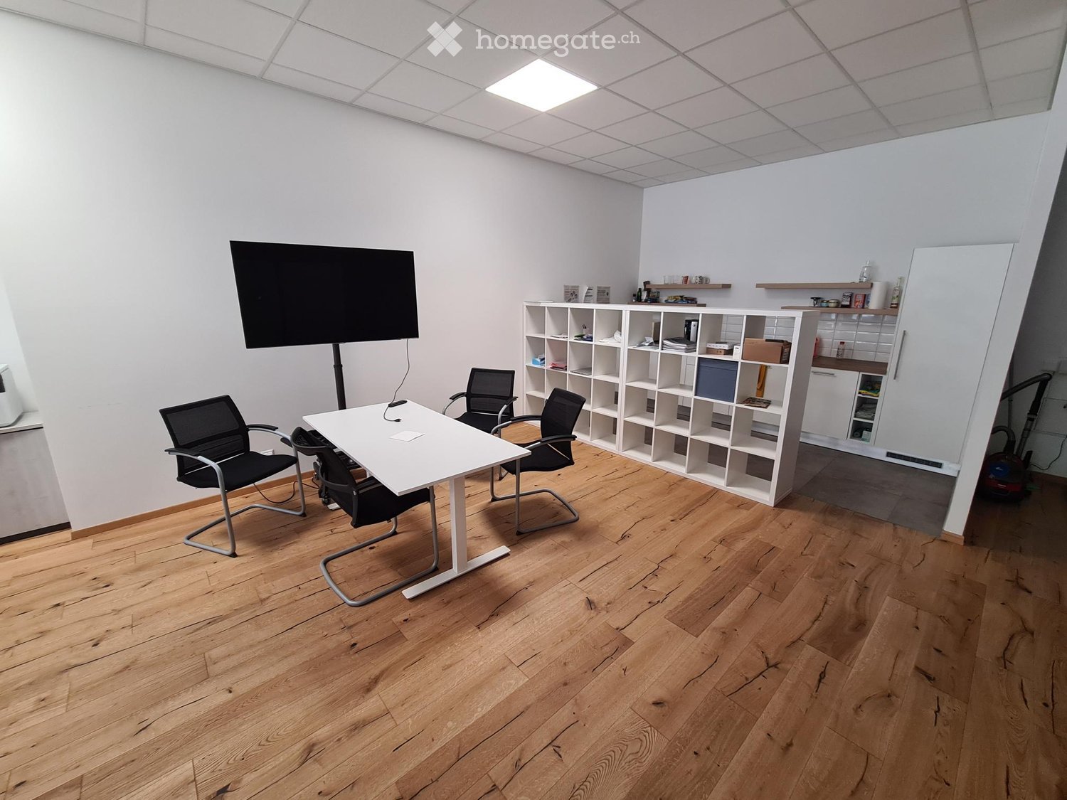 Office space with wooden flooring, white table, four black chairs, white shelving unit, and a large screen on a stand.
