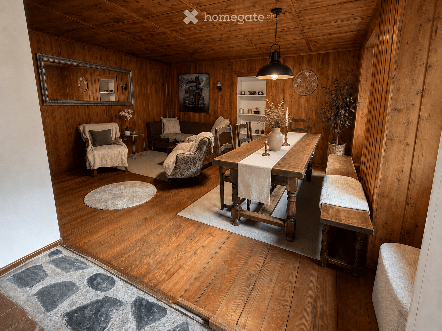 Cozy living and dining area with wooden walls and ceiling, featuring a sofa, chairs, a dining table with bench seating, a vase with flowers, a mirror, and decorative artwork.